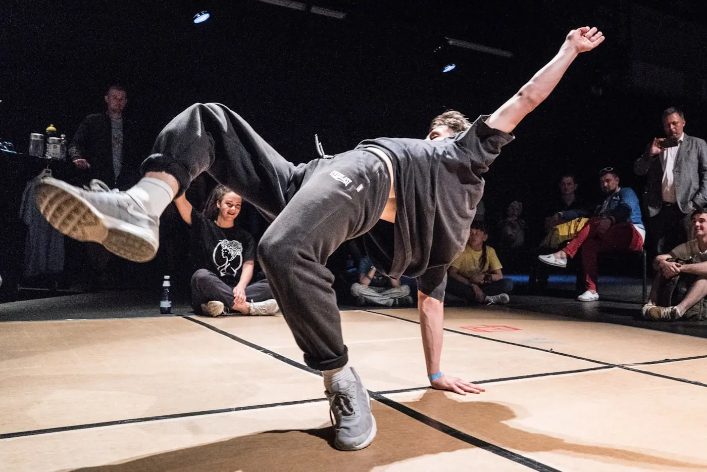A dancer holding a freeze inside a circle of people on a wooden floor.