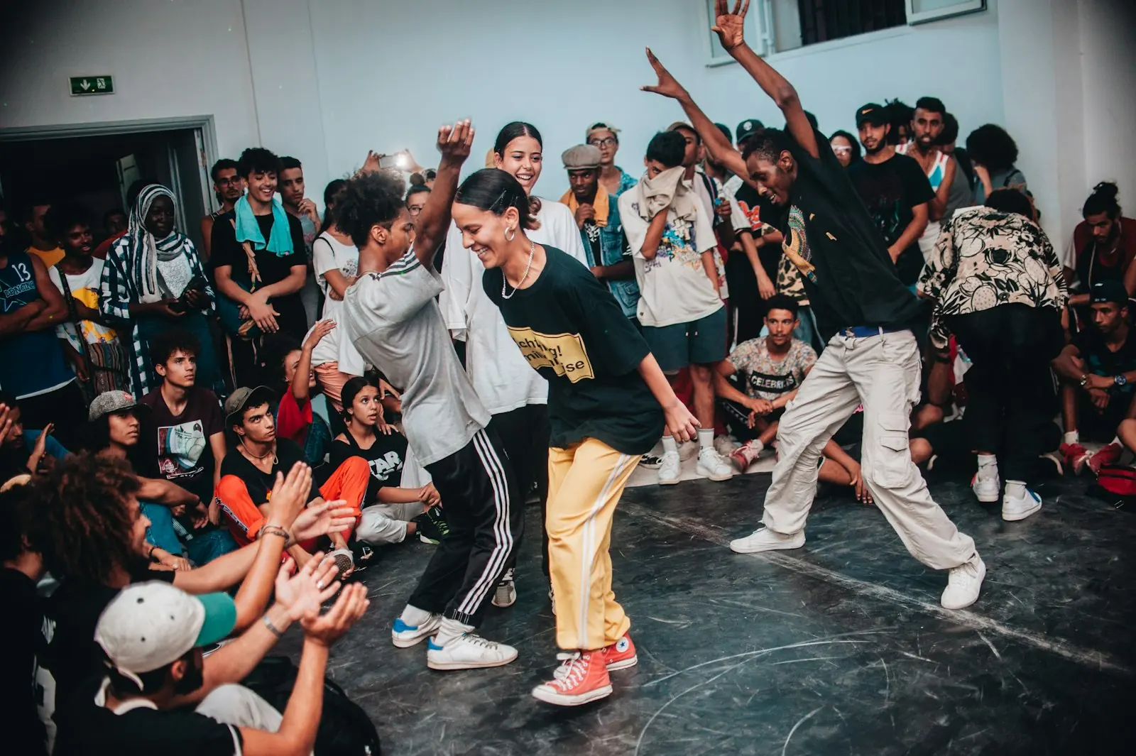A circle of dancers watching one person in the middle, arms up, room full.